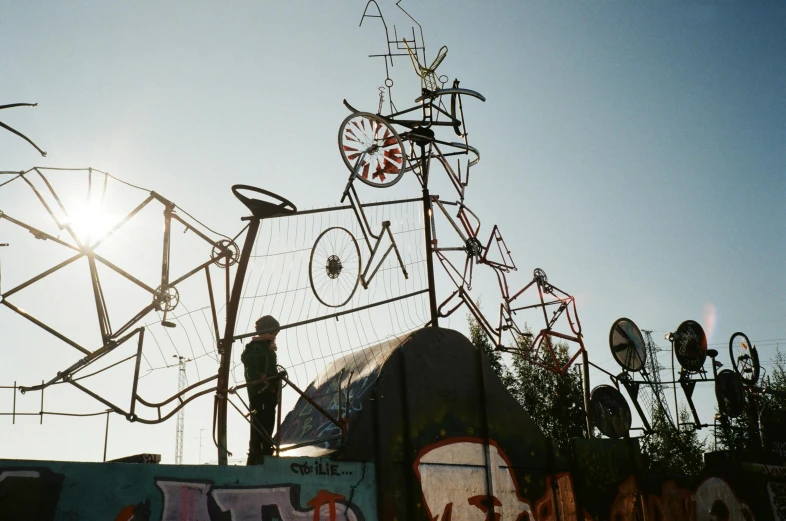 a man standing on top of a graffiti covered wall, an abstract sculpture, by Attila Meszlenyi, unsplash, kinetic art, machine garden, standing astride a gate, complex contraption, early evening