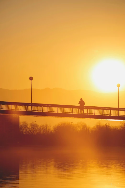 a couple of people that are standing on a bridge, pexels contest winner, romanticism, bright yellow and red sun, albuquerque, morning haze, lone person in the distance