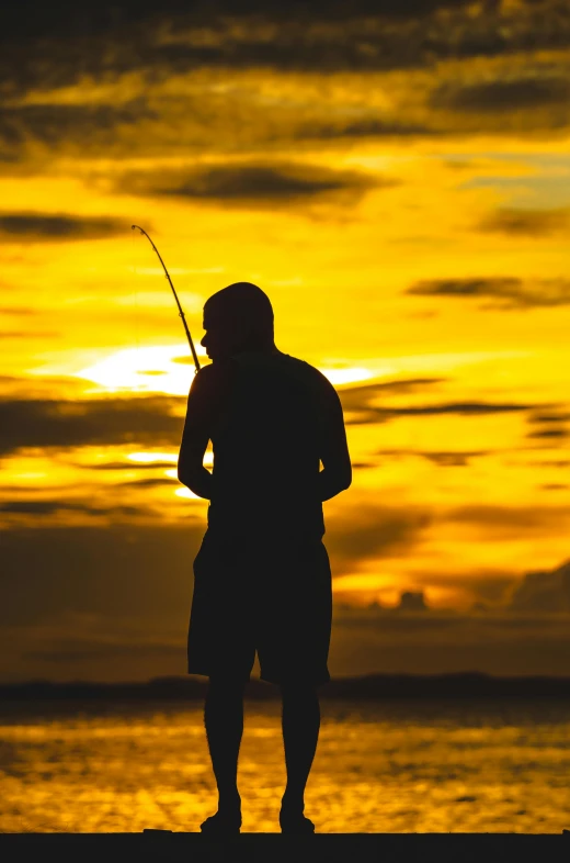 a man standing on top of a beach holding a fishing pole, during a sunset