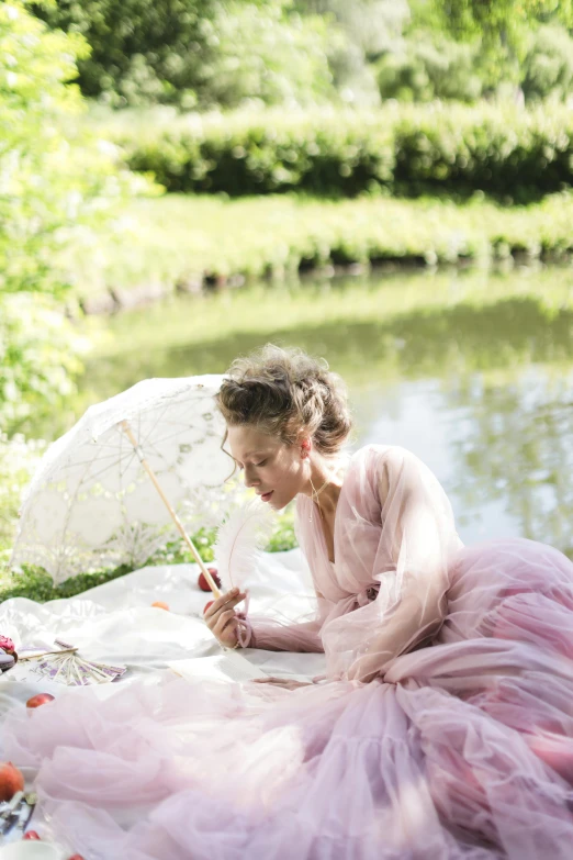 a woman in a pink dress sitting on a blanket with an umbrella, inspired by Konstantin Somov, unsplash, romanticism, garden pond scene, tulle and lace, nathalie emmanuel, behind the scenes