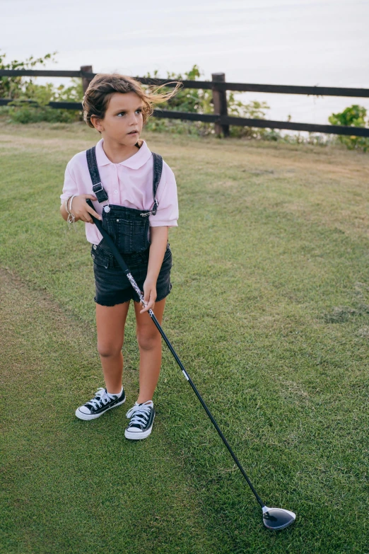 a little girl standing on top of a green field holding a golf club, inspired by Shirley Teed, unsplash, happening, suspenders, looking down on the camera, wearing polo shirt, embarrassing