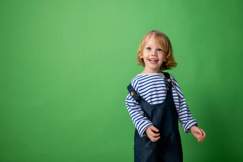 a little girl standing in front of a green wall, by Helen Stevenson, unsplash, blue overalls, wearing stripe shirt, studio shoot, while smiling for a photograph