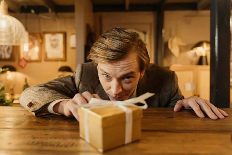 a man leaning over a table with a present in front of him, by Alice Mason, pexels contest winner, wes anderson film, looking surprised, eleventh doctor, mark e smith