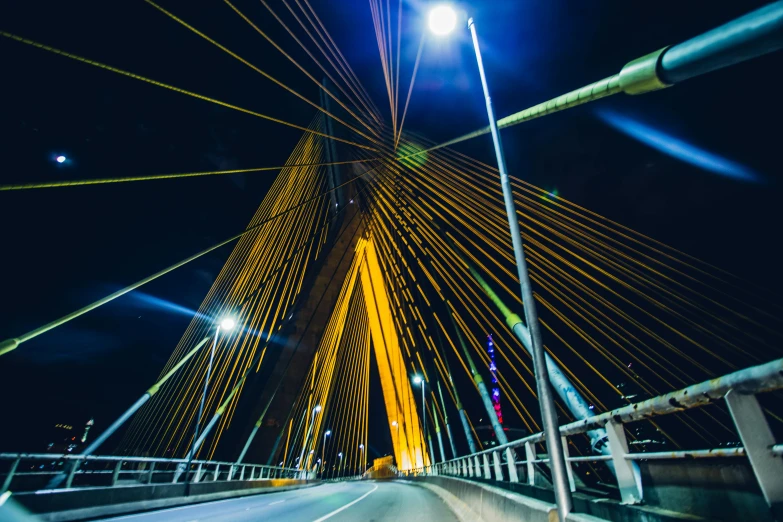 a car driving across a bridge at night, by Ibrahim Kodra, unsplash contest winner, hurufiyya, gold cables, blue and yellow lighting, flawless structure, wideangle