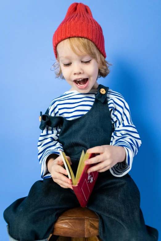 a little boy sitting on a stool reading a book, by Paul Bird, pexels, mannerism, wearing a french beret, blue overalls, mini model, giggling