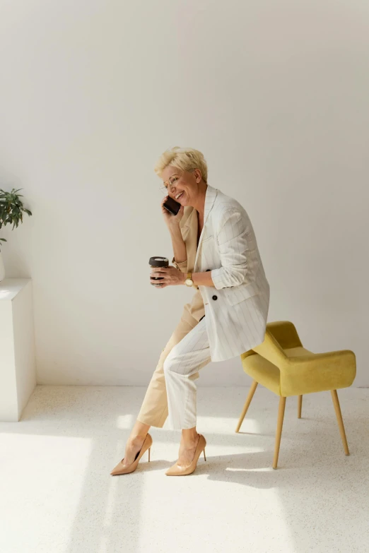 a woman sitting on a chair talking on a cell phone, trending on pexels, minimalism, tan suit, older woman, white, morning coffee