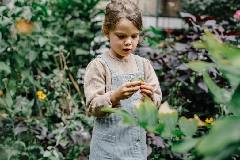 a little girl standing in a garden looking at a flower, inspired by Elsa Beskow, pexels contest winner, wearing dirty overalls, grey, foliage clothing, press shot