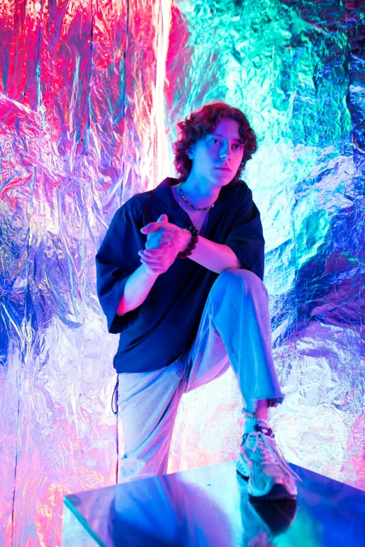 a man sitting on top of a table next to a waterfall, an album cover, unsplash, magical realism, timothee chalamet, iridescent neon, distorted pose, foil