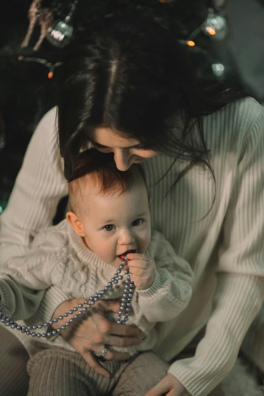 a woman holding a baby in front of a christmas tree, pexels contest winner, symbolism, neck chains, avatar image, grey, cords