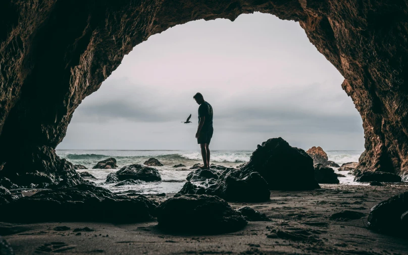 a man standing in a cave looking out at the ocean, by Matt Cavotta, unsplash contest winner, hollister ranch, slight overcast, a photo of a man, a person standing in front of a