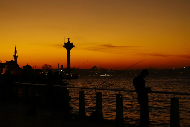 a man standing on top of a pier next to a body of water, a picture, by Fikret Muallâ Saygı, hurufiyya, city sunset night, silhouette :7, fisherman, istanbul