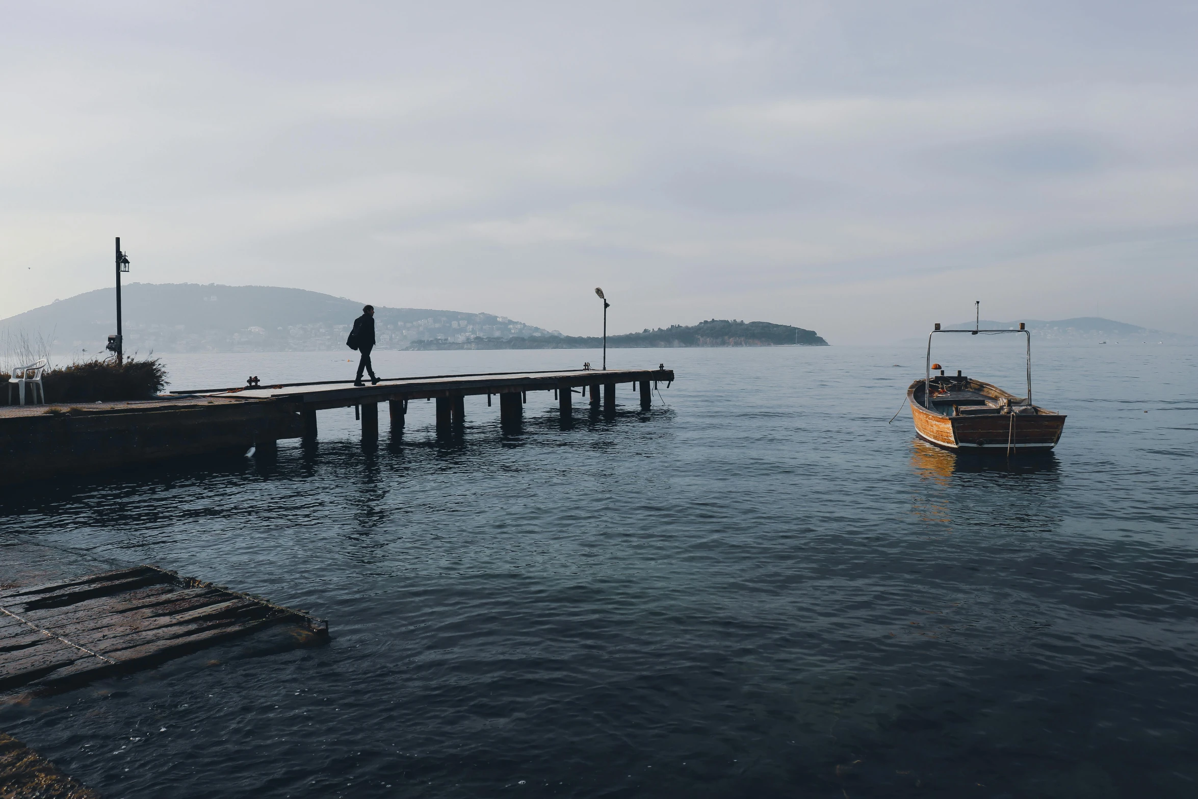 a person standing on a dock next to a boat, by Niyazi Selimoglu, serene overcast atmosphere, people walking around, brown, unedited