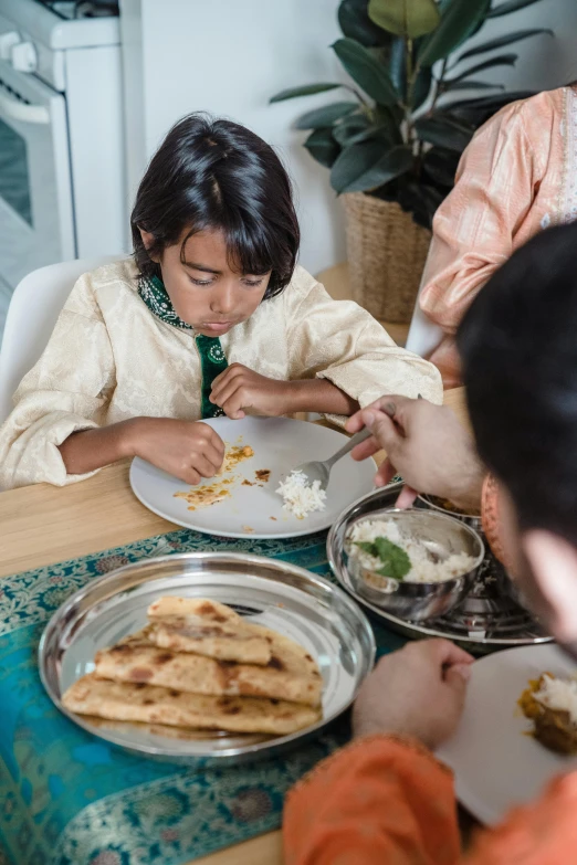 a group of people sitting around a table eating food, by Maryam Hashemi, pexels contest winner, hurufiyya, little kid, holding a kitchen knife, hindu aesthetic, over a dish and over a table
