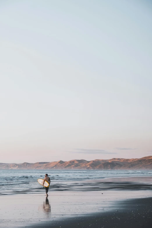 a person walking on a beach with a surfboard, by Peter Churcher, unsplash contest winner, new zeeland, hills, late summer evening, crisp clean shapes