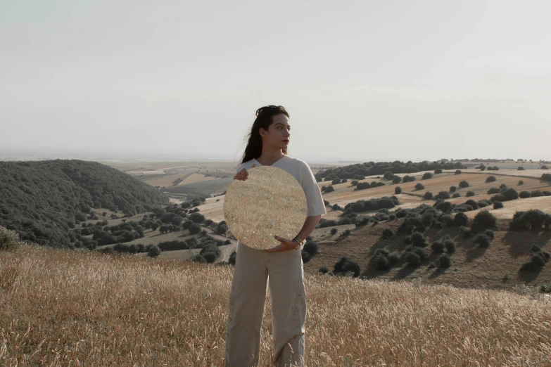 a woman standing on top of a grass covered hillside, an album cover, pexels contest winner, land art, holding a shield, cyprus, round-cropped, white marble and gold