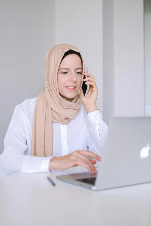 a woman sitting in front of a laptop talking on a cell phone, inspired by Maryam Hashemi, trending on pexels, hurufiyya, white scarf, wearing business casual dress, white helmet, malaysian