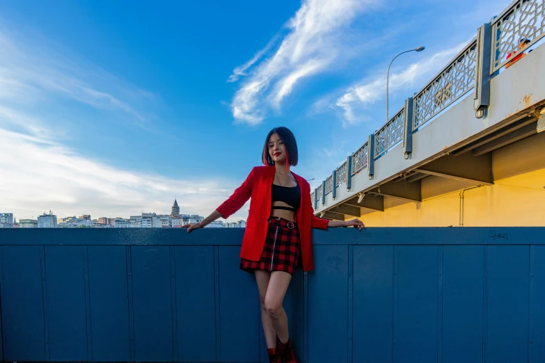 a woman standing on top of a blue fence, by Julia Pishtar, pexels contest winner, short skirt and a long jacket, red and black suit, standing on a bridge, russian and japanese mix