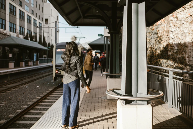 a woman waiting for a train at a train station, by Carey Morris, trending on unsplash, happening, man and woman walking together, seattle, standing with her back to us, bus stop