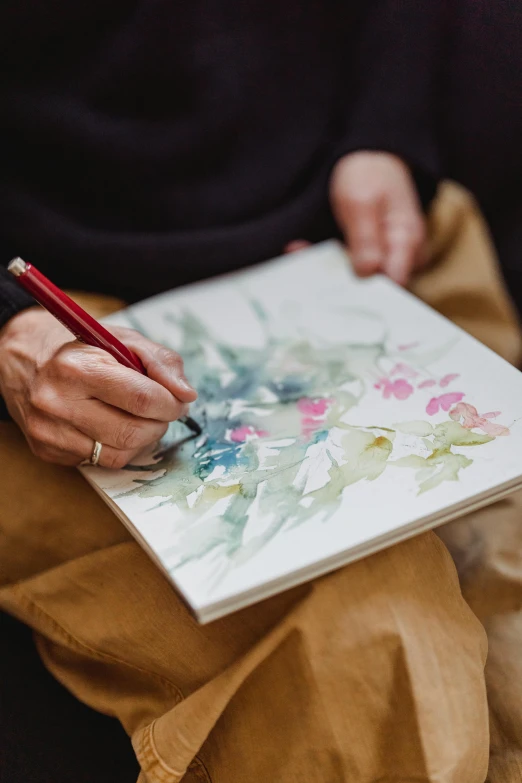 a person holding a pen and drawing on a piece of paper, a watercolor painting, inspired by senior artist, trending on unsplash, art book, botanic watercolors, on canvas, ((water color))