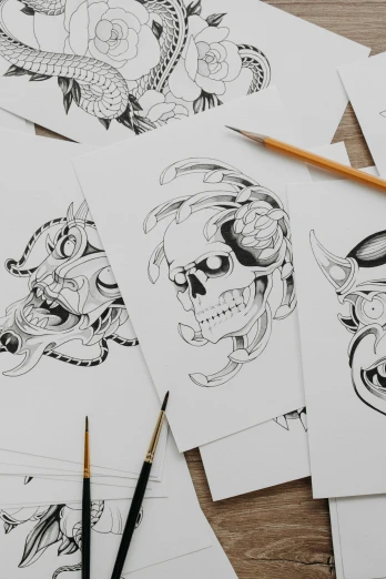 a bunch of drawings sitting on top of a wooden table, an ink drawing, inspired by Kawanabe Kyōsai, trending on pexels, skull tattoo, on white paper, symmetrical artwork, professional illustration