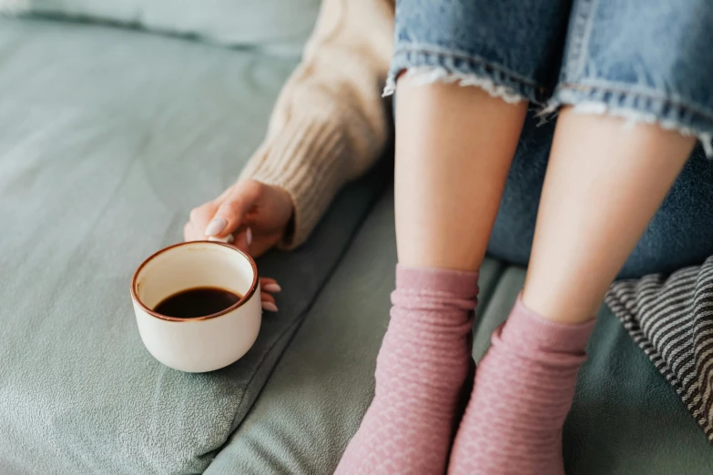 a woman sitting on a couch holding a cup of coffee, inspired by Elsa Bleda, trending on pexels, feet on the ground, sustainable materials, loputyn and matcha, girl with brown hair