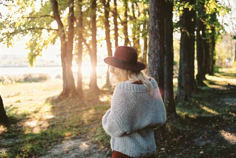 a woman in a hat is standing in the woods, trending on pexels, wearing sweater, sunny afternoon, facing away, softly - lit