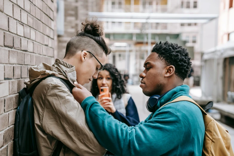 a couple of people standing next to a brick wall, trending on pexels, happening, black teenage boy, woman holding another woman, bullying, an olive skinned