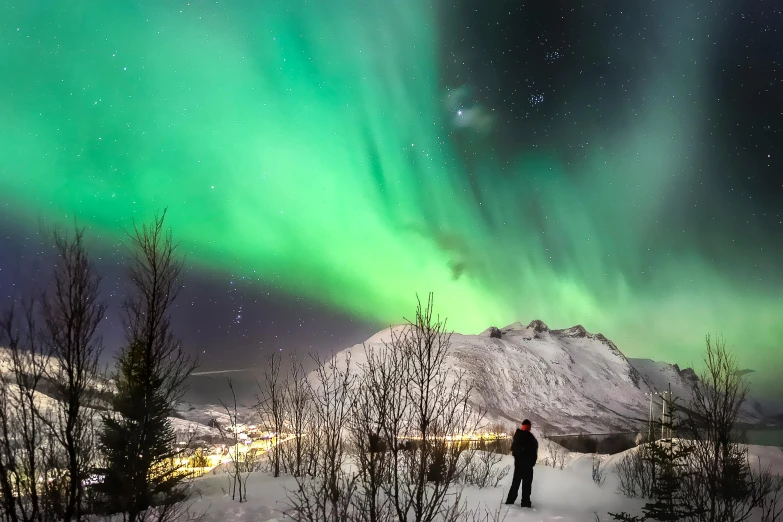 a man standing on top of a snow covered slope, by Terese Nielsen, pexels contest winner, visual art, northen lights background, distant mountains lights photo, nat geo, grey