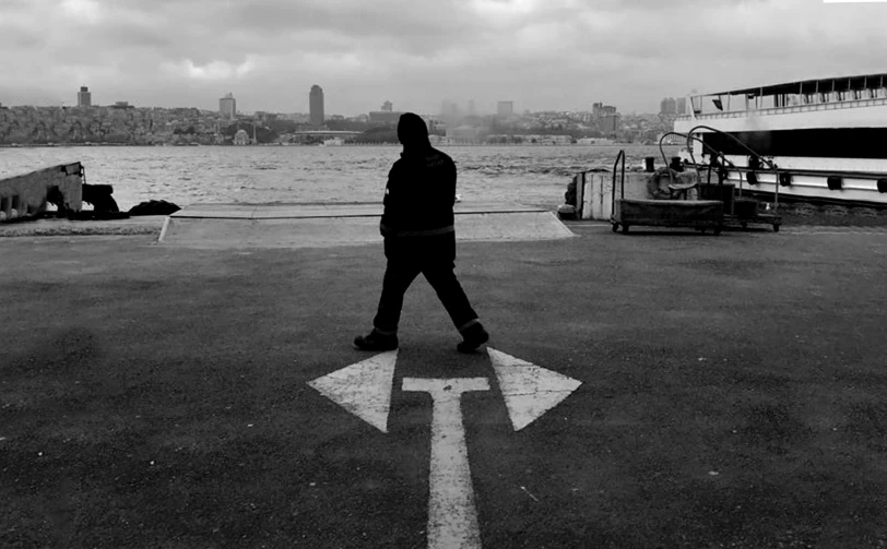 a man walking across a parking lot next to a body of water, a black and white photo, arrow, new york harbour, istanbul, facebook photo