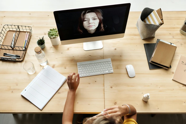 a woman sitting at a desk in front of a computer, trending on pexels, video art, high - angle view, face showing, on a wooden table, schools