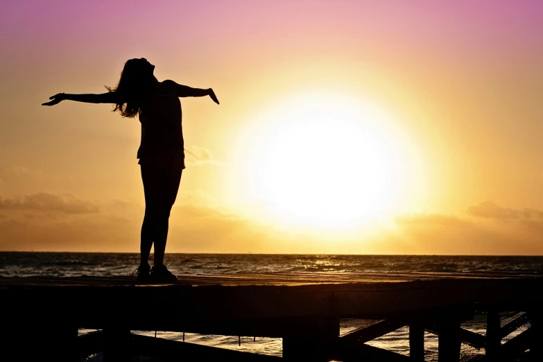 a woman standing on a pier at sunset, pexels contest winner, arms out, relaxing, silhouette :7, happy girl