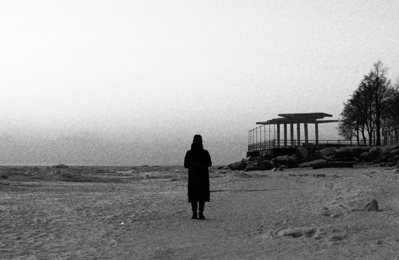 a black and white photo of a person walking on a beach, a black and white photo, inspired by Sergio Larraín, unsplash, conceptual art, woman in black robes, silent hill aesthetic, cold winter, still from a music video