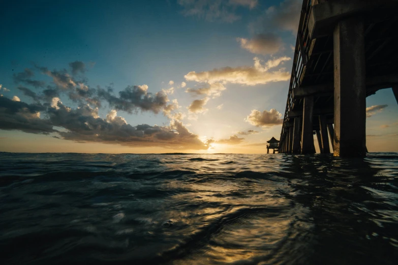 a person riding a surfboard under a bridge, by Niko Henrichon, unsplash contest winner, renaissance, sunset with cloudy skies, ferrofluid oceans, hd wallpaper, near a jetty