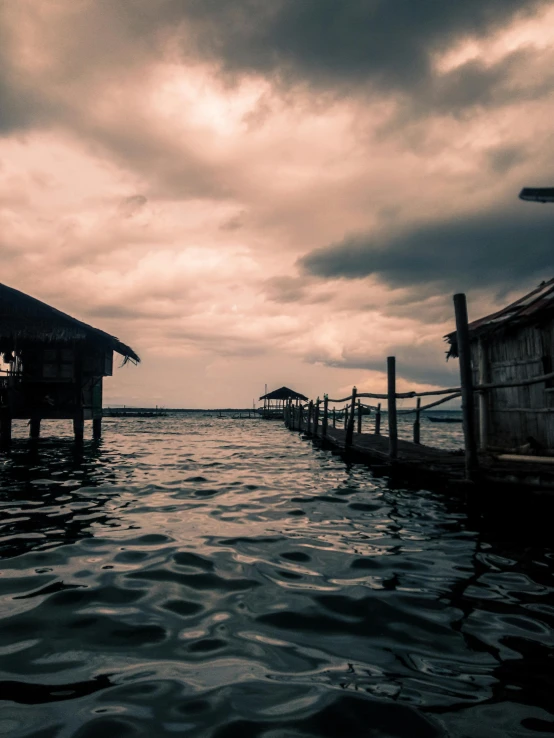 a pier next to a body of water under a cloudy sky, a picture, pexels contest winner, sumatraism, houses on stilts, album cover, it's getting dark, calmly conversing 8k