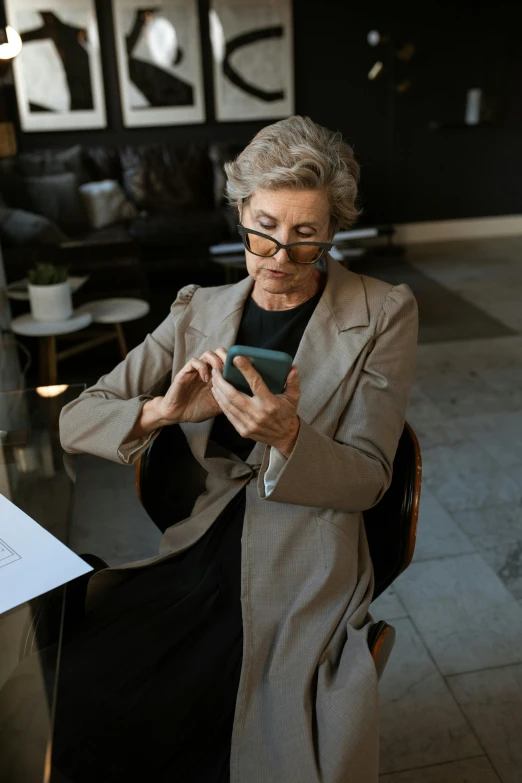 a woman sitting in a chair looking at her cell phone, a portrait, trending on pexels, old lady cyborg merchant, wearing a worn out brown suit, wearing black rimmed glasses, architect