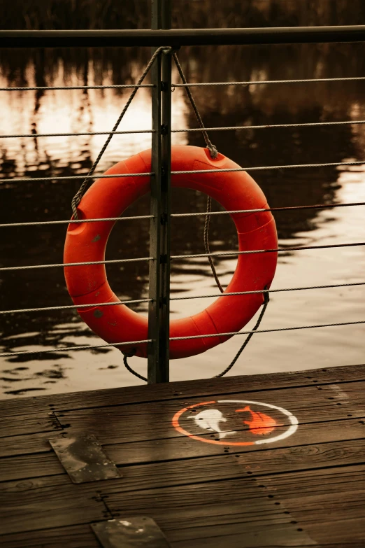 a life preserver sitting on a dock next to a body of water, by Eglon van der Neer, pexels contest winner, symbolism, orange safety labels, red river, softly lit, circular shape