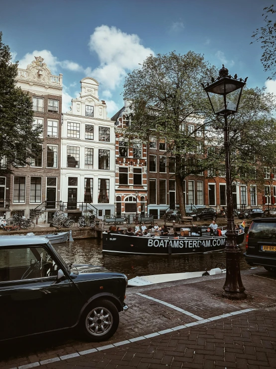 a black car driving down a street next to tall buildings, by Jacob Koninck, dutch houses along a river, set photo, 🚿🗝📝