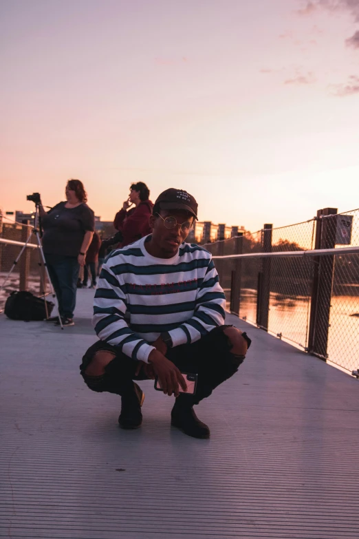 a man squatting down in front of a body of water, an album cover, pexels contest winner, city sunset, playboi carti portrait, vsco, standing on a bridge