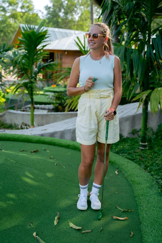 a woman standing on top of a green putting green, a portrait, unsplash, wearing a camisole and shorts, a blond, tropical setting, sports setting