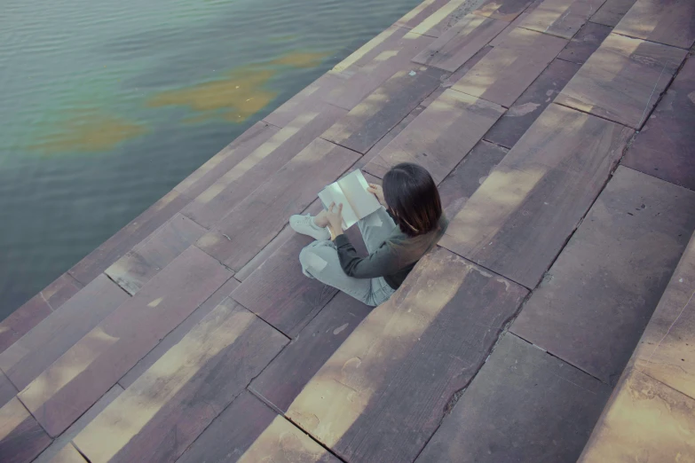 a woman sitting on steps next to a body of water, inspired by Oleg Oprisco, pexels contest winner, hyperrealism, reading the book about love, top down perspecrive, grey, video still