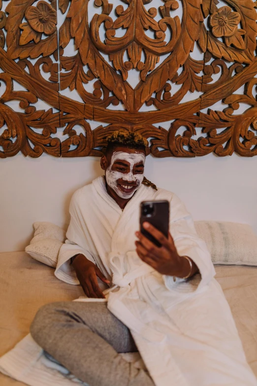 a man sitting on a bed holding a cell phone, white facepaint, bali, skincare, instagram picture