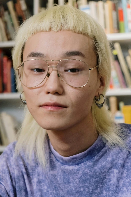 a man with blonde hair and glasses sitting in front of a bookshelf, inspired by jeonseok lee, featured on reddit, rococo, huge earrings and queer make up, closeup headshot, slide show, asian descent