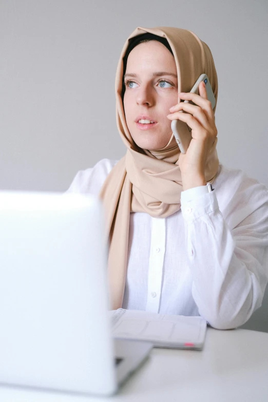 a woman sitting at a table talking on a cell phone, inspired by Maryam Hashemi, trending on pexels, hurufiyya, white hijab, 15081959 21121991 01012000 4k, female in office dress, focused stare