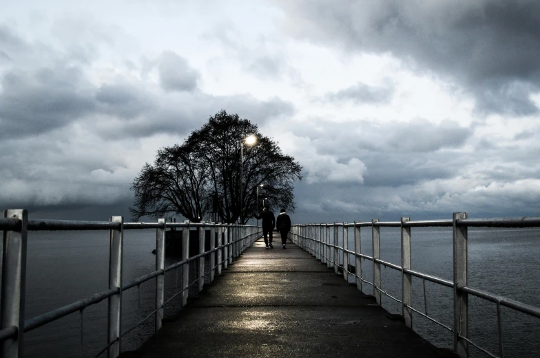 a pier next to a body of water under a cloudy sky, by Lucia Peka, pexels contest winner, romanticism, people walking around, it's getting dark, two people, gloomy weather. high quality