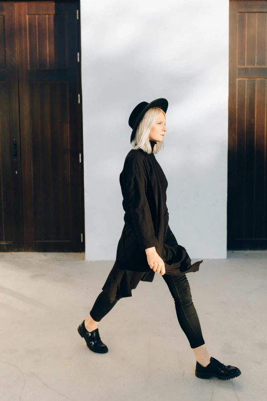 a woman walking in front of a white wall, by Sara Saftleven, trending on unsplash, casual black clothing, black hat, long tunic, concert