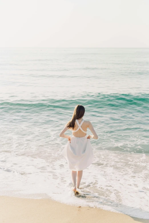 a woman standing on top of a beach next to the ocean, open back dress, cresting waves and seafoam, wearing white clothes, sun yunjoo