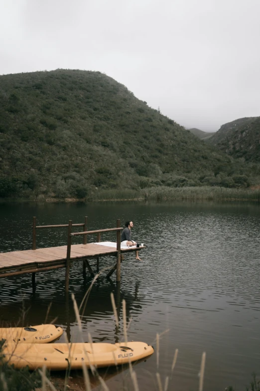 a man sitting on a dock next to a body of water, a picture, by Elsa Bleda, unsplash, hurufiyya, south african coast, 2 5 6 x 2 5 6 pixels, river and trees and hills, female floating