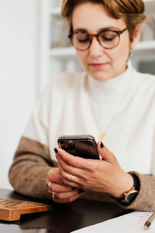 a woman sitting at a table looking at her cell phone, trending on pexels, wearing a white sweater, wearing black rimmed glasses, corporate phone app icon, wearing a brown