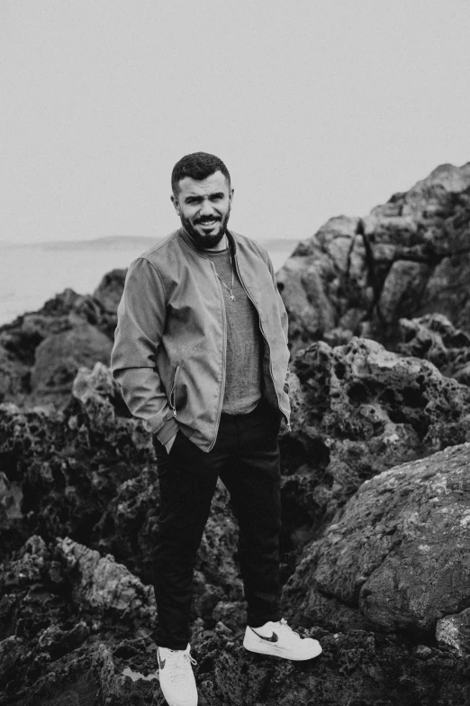 a man standing on top of a rocky beach, a black and white photo, charli bowater and artgeem, headshot profile picture, smiling male, demna gvasalia