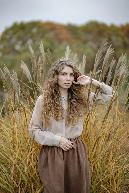 a woman standing in a field of tall grass, by irakli nadar, trending on pexels, renaissance, brown fluffy hair, autum, pale skin curly blond hair, brown shirt